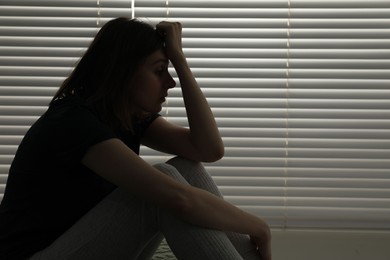 Sad young woman near window indoors, space for text