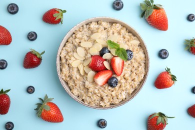 Photo of Tasty oatmeal with strawberries, blueberries and almond flakes in bowl surrounded by fresh berries on light blue background, flat lay