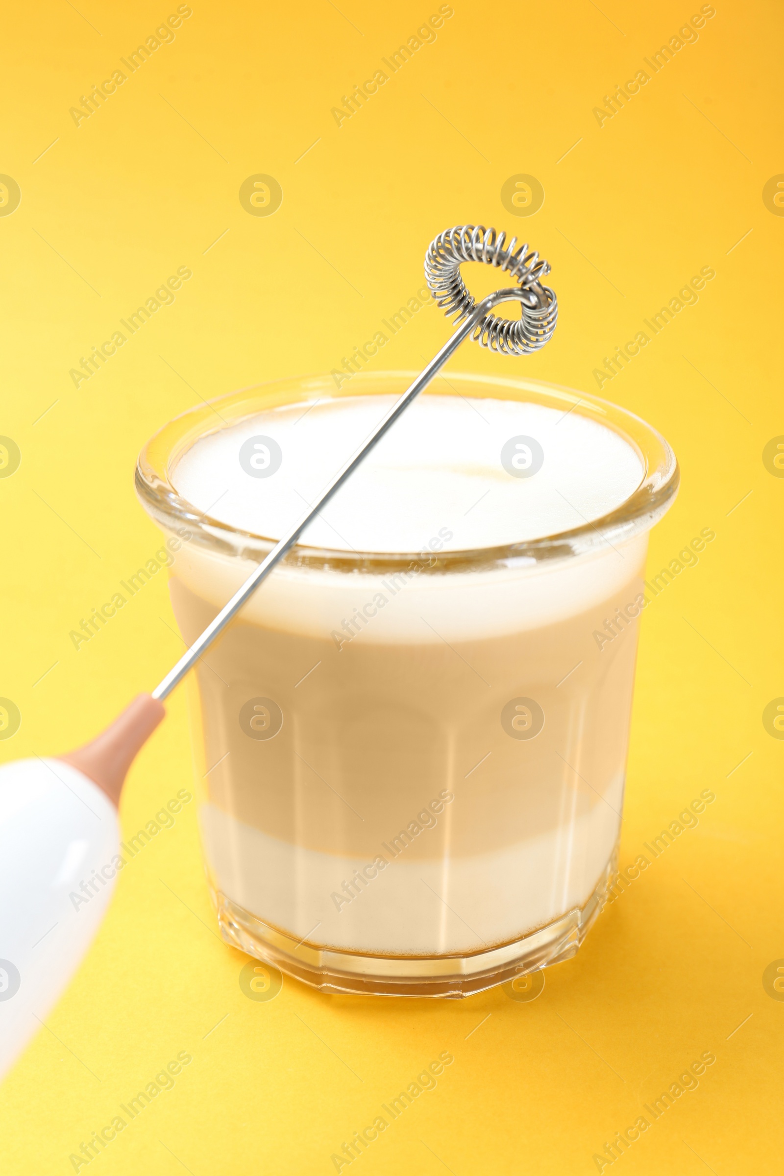 Photo of Mini mixer (milk frother) and tasty cappuccino in glass on yellow background