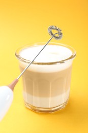 Mini mixer (milk frother) and tasty cappuccino in glass on yellow background