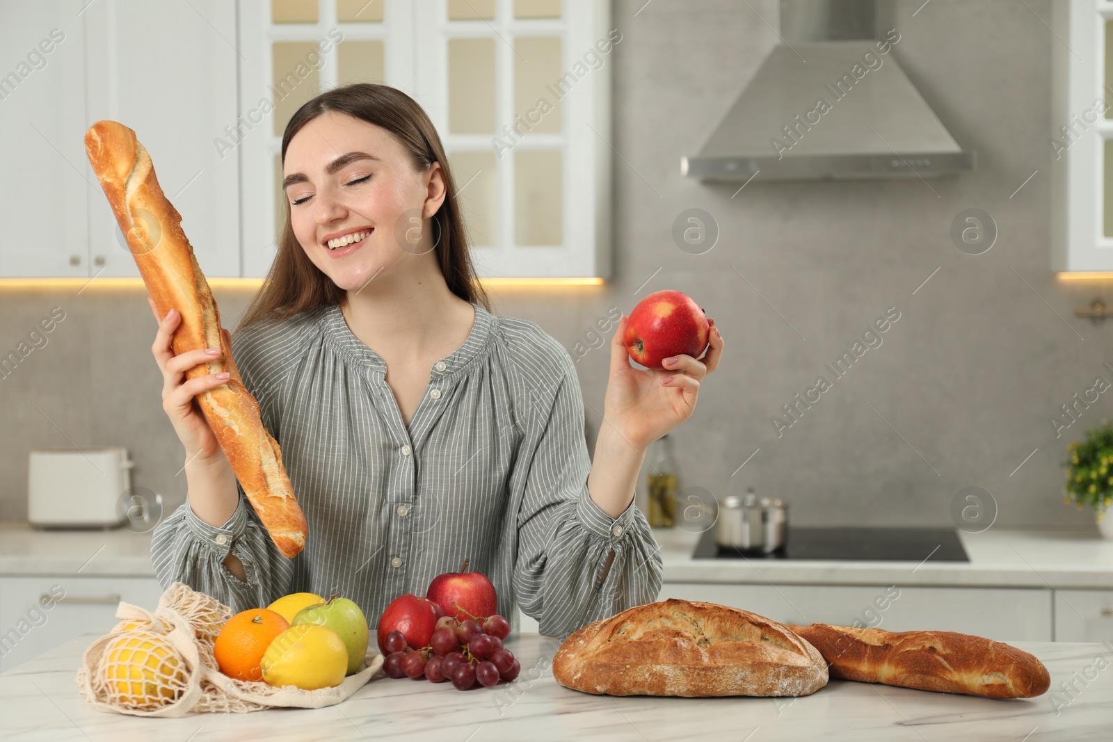 Photo of Woman with baguette, apple and string bag of fresh fruits at light marble table in kitchen