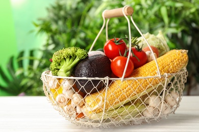 Photo of Fresh vegetables in metal basket on white wooden table against blurred green background