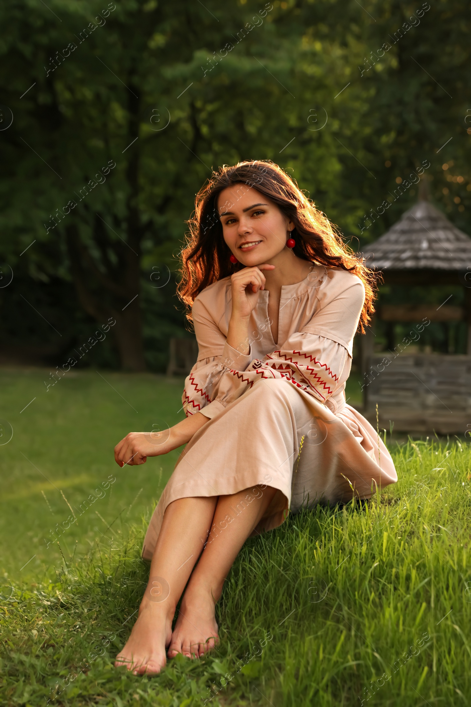 Photo of Beautiful woman in embroidered dress sitting on green grass in village. Ukrainian national clothes