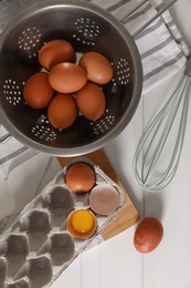 Fresh raw chicken eggs with colander, napkin and whisk on white wooden table, flat lay