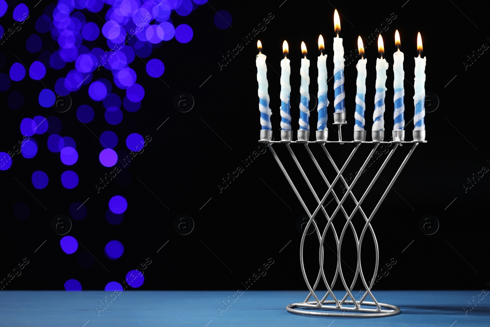 Photo of Hanukkah celebration. Menorah with burning candles on blue table against dark background with blurred lights, space for text