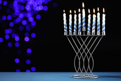 Photo of Hanukkah celebration. Menorah with burning candles on blue table against dark background with blurred lights, space for text