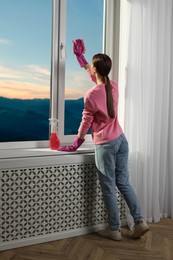 Photo of Young woman cleaning window glass with rag at home, back view