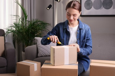 Photo of Young woman using utility knife to open parcel at home