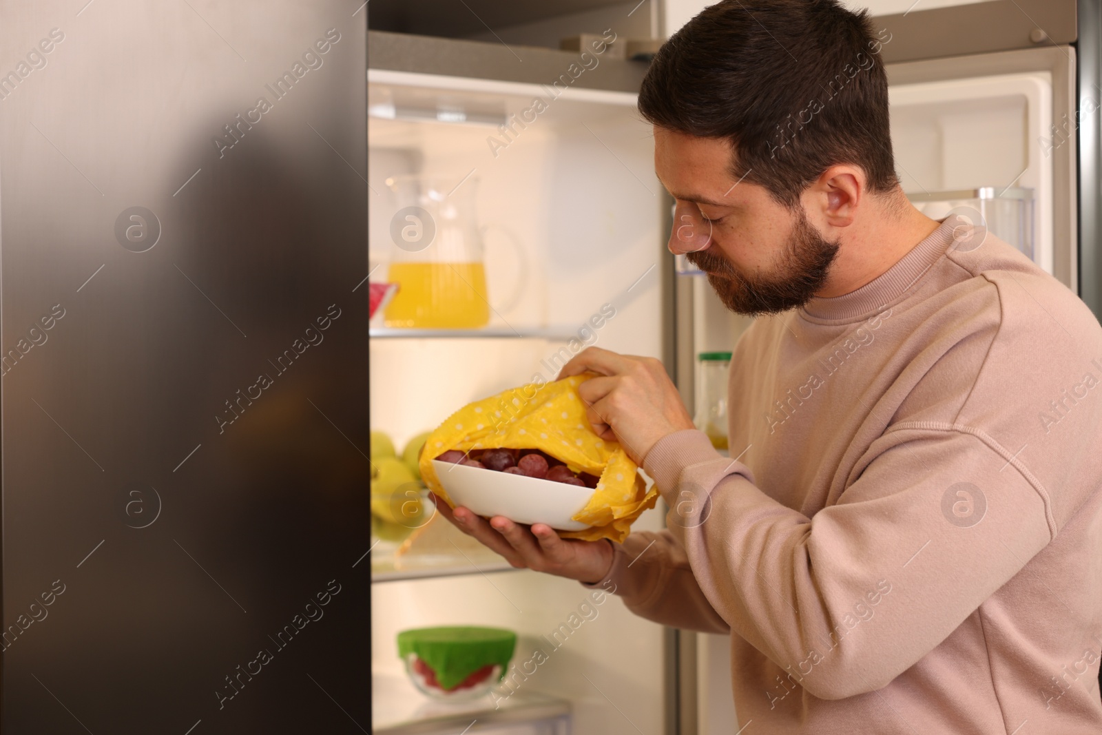 Photo of Man putting bowl covered with beeswax food wrap into refrigerator indoors