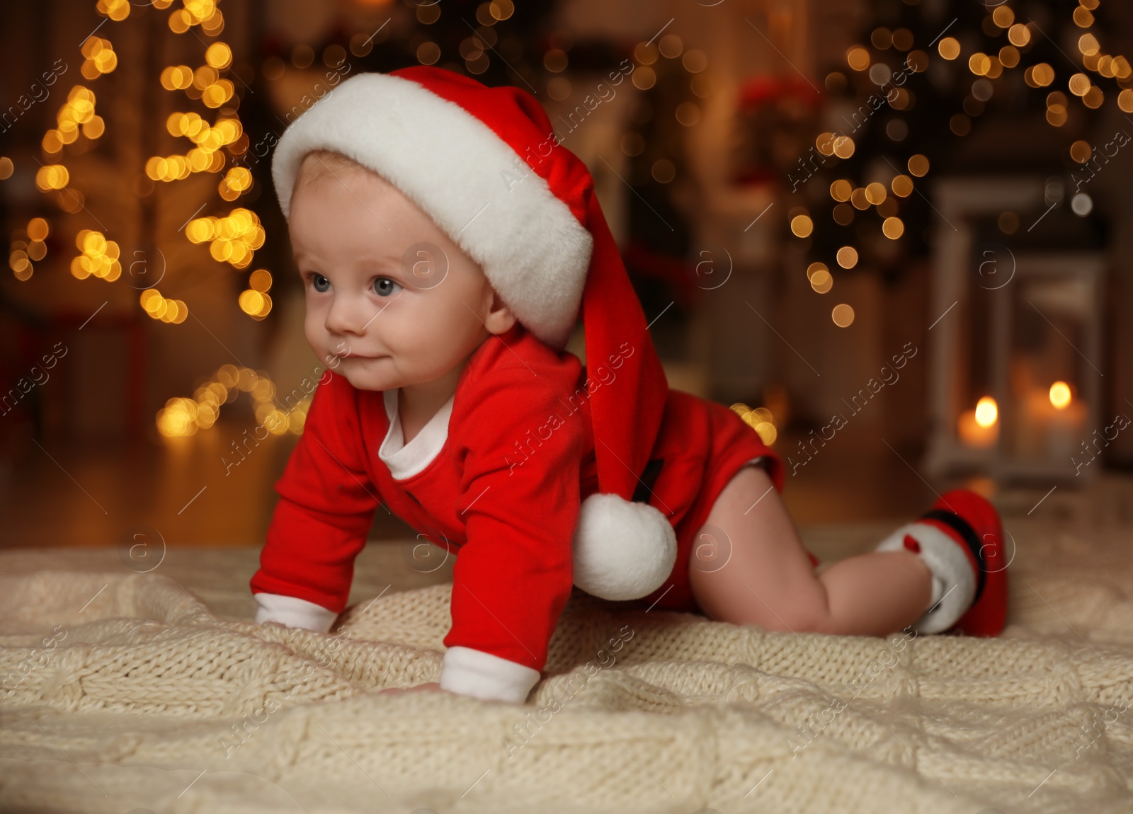 Photo of Cute little baby in Santa Claus suit on blanket against blurred festive lights. Christmas celebration