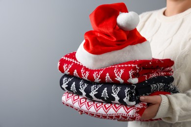 Photo of Woman holding stack of different Christmas sweaters and Santa hat on grey background, closeup. Space for text