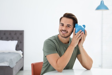 Young man with piggy bank at table indoors. Space for text