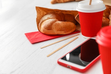 Photo of Cup of coffee, bun in paper bag and smartphone on wooden table