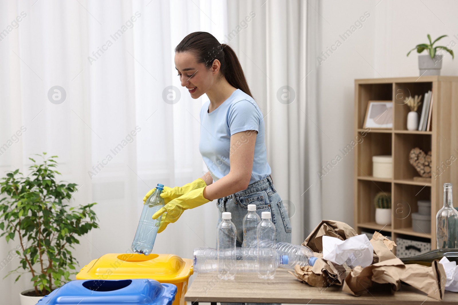 Photo of Garbage sorting. Smiling woman throwing plastic bottle into trash bin in room