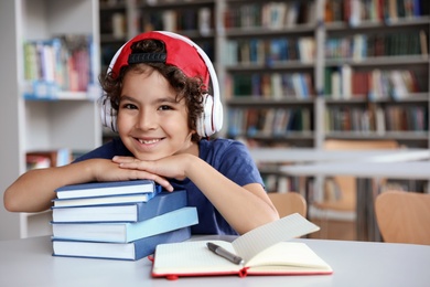 Cute little boy with headphones and books at table in library reading room