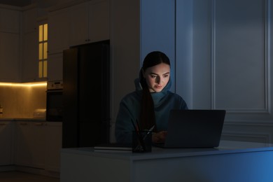 Young woman using laptop at table in kitchen at night