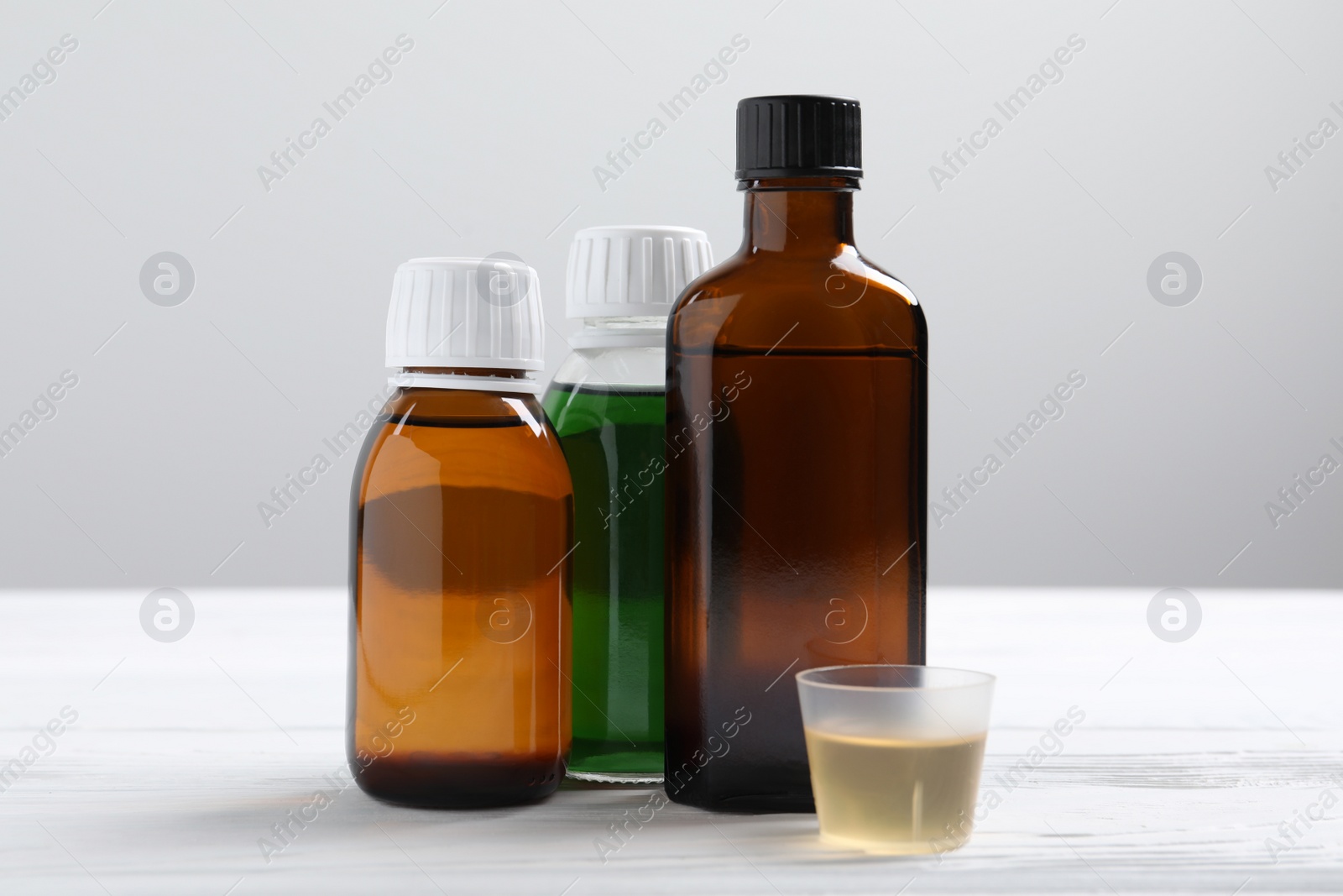 Photo of Bottles of syrup and measuring cup on white table against light grey background. Cold medicine