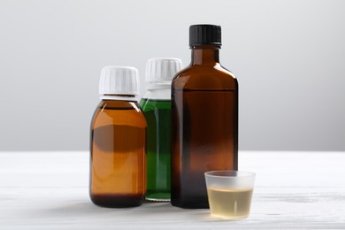 Photo of Bottles of syrup and measuring cup on white table against light grey background. Cold medicine