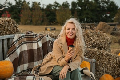 Photo of Beautiful woman with cup of hot drink sitting on wooden bench near pumpkins and hay bales outdoors. Autumn season