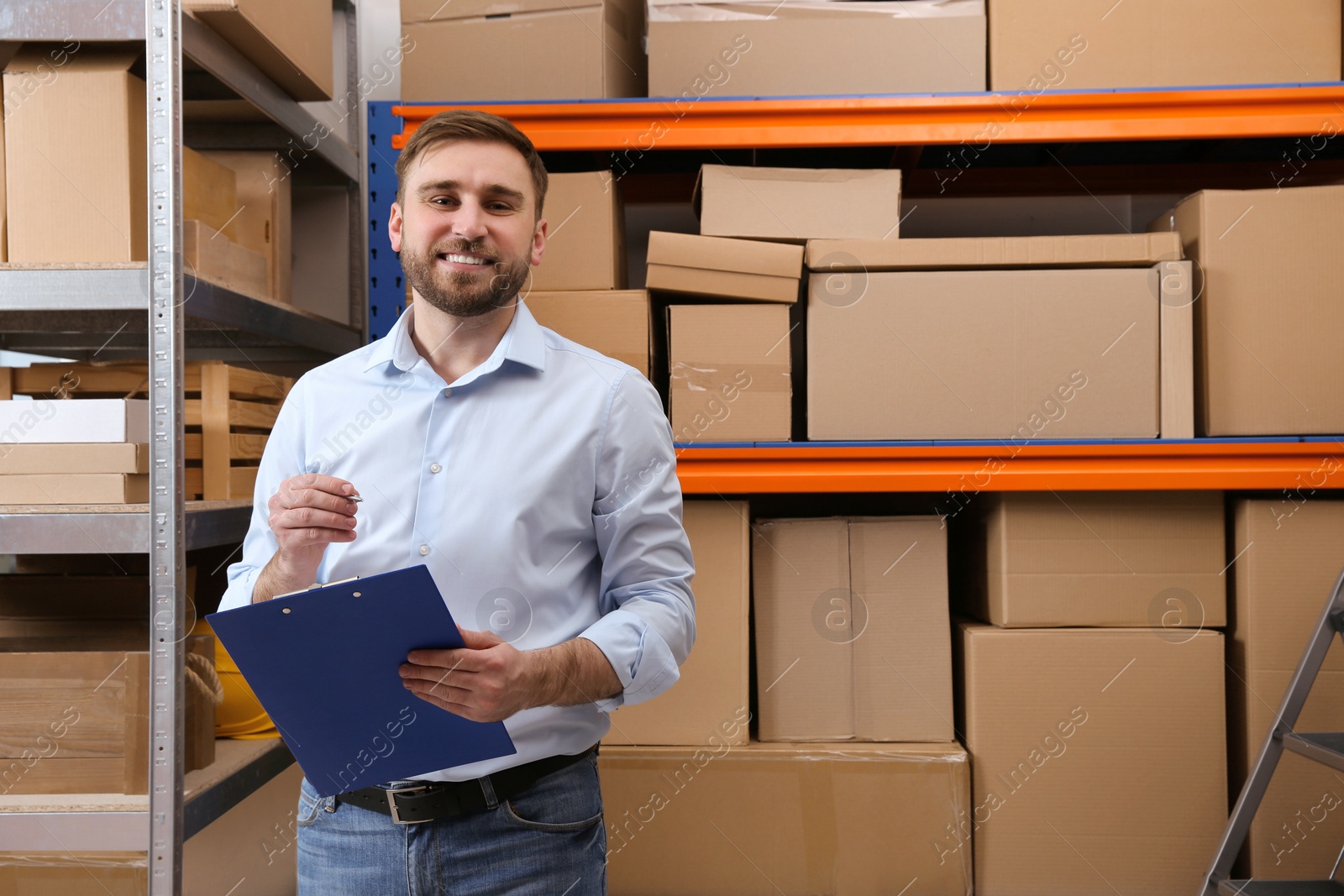 Photo of Young businessman with clipboard near rack of cardboard boxes at warehouse