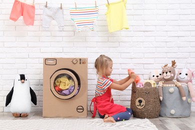 Photo of Little girl putting laundry into basket near toy cardboard washing machine indoors