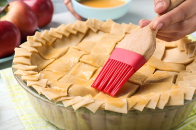 Photo of Woman spreading egg yolk onto raw apple pie at white wooden table, closeup