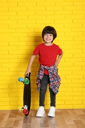 Photo of Cute little boy with skateboard near yellow brick wall indoors