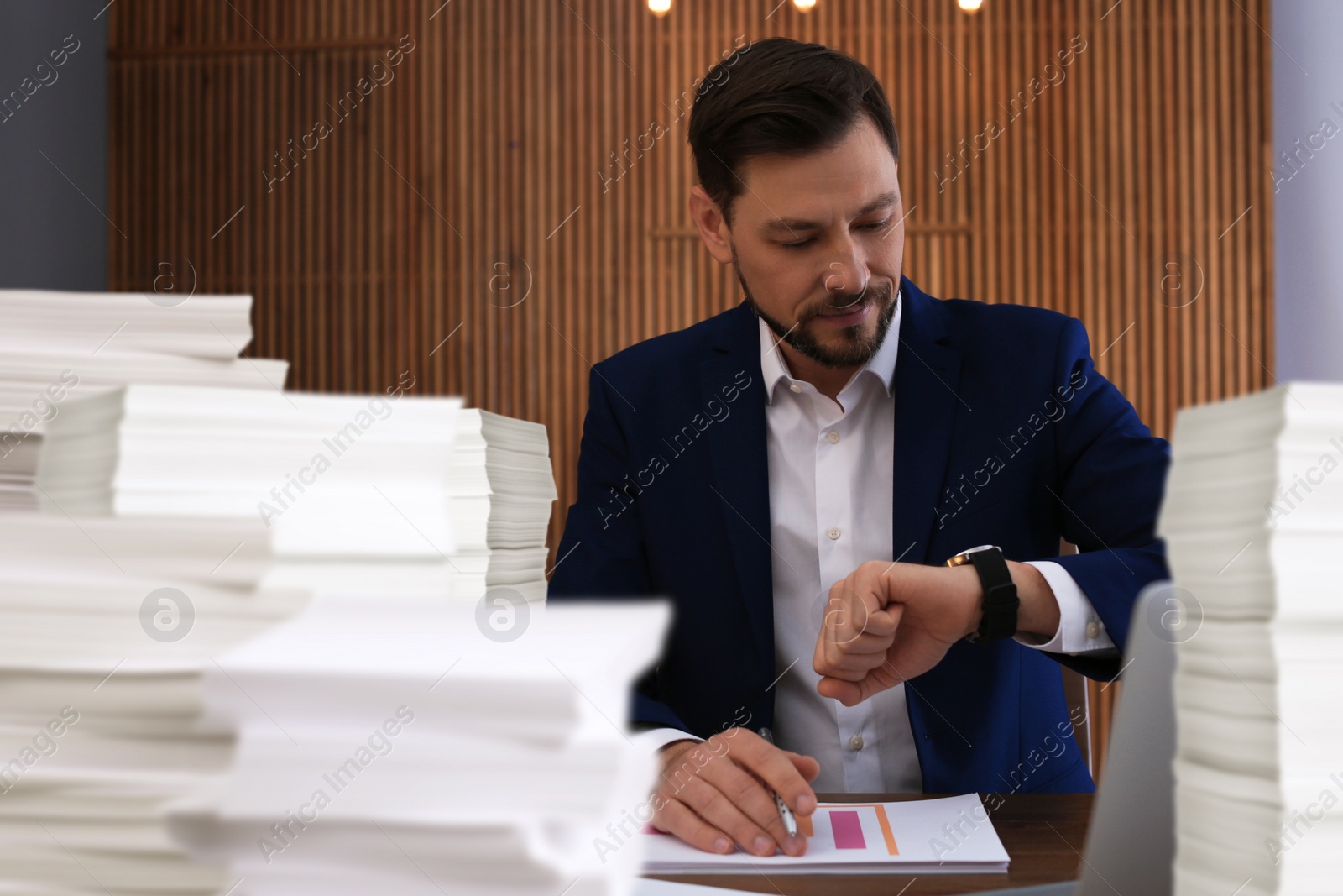 Image of Deadline concept. Busy man looking at wristwatch in office. His workplace with lots of documents