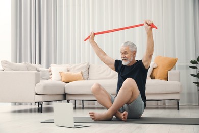 Photo of Senior man doing exercise with fitness elastic band near laptop on mat at home