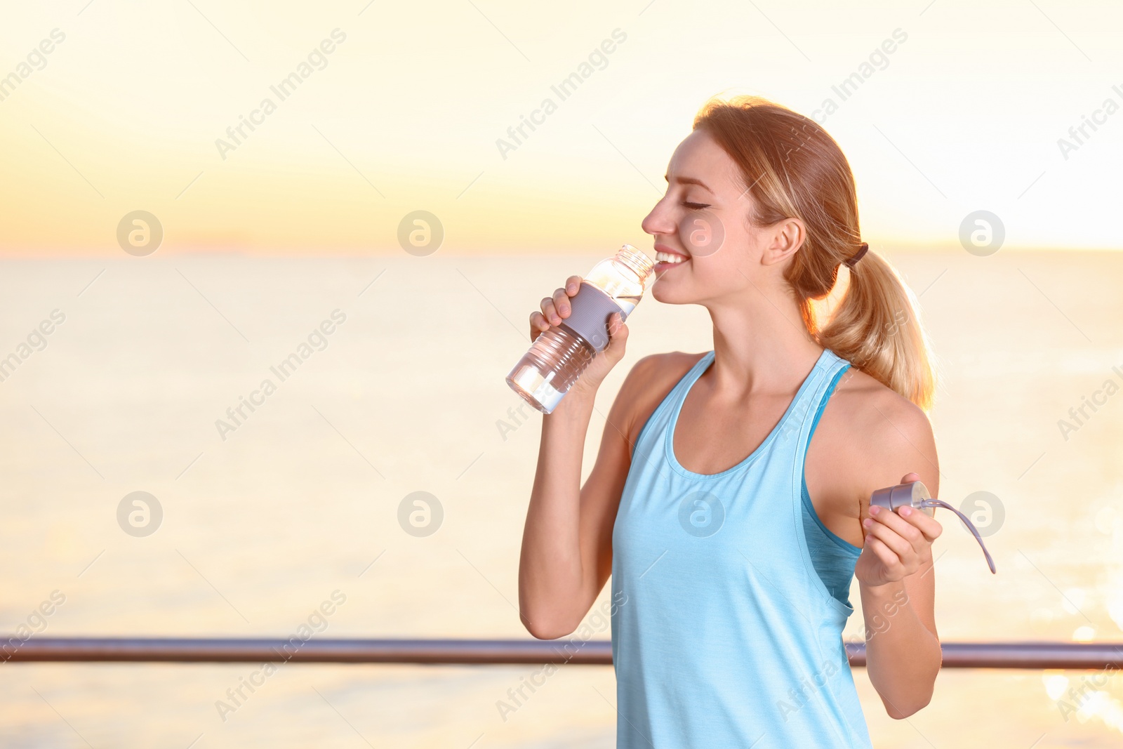 Photo of Young woman drinking water from bottle after fitness exercises on pier in morning