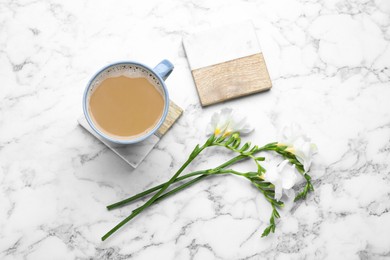 Photo of Mug of coffee, stylish cup coasters and beautiful freesia flowers on white marble table, flat lay