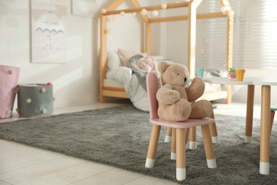 Photo of Small table, chair with bunny ears and teddy bear in children's bedroom interior