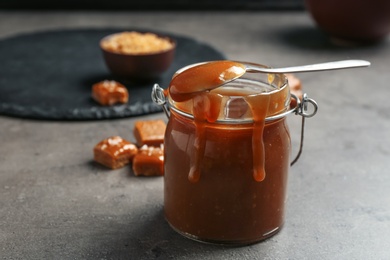 Photo of Jar with tasty caramel sauce and spoon on grey table