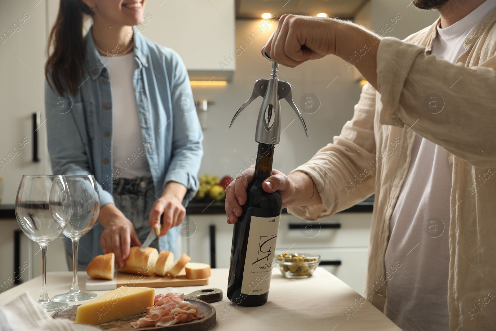 Photo of Man opening wine bottle with corkscrew while smiling woman cutting bread at table indoors, closeup