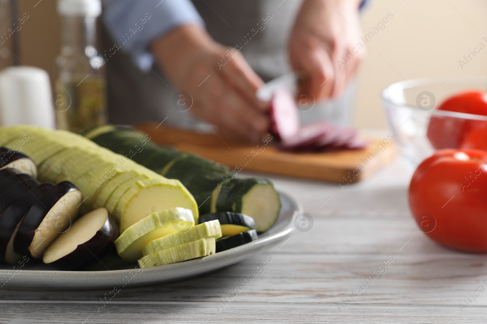 Photo of Cooking delicious ratatouille. Plate with fresh vegetables and woman cutting onion at white wooden table, closeup. Space for text