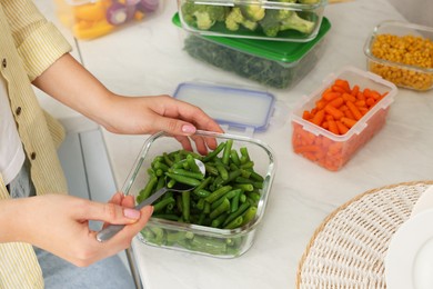 Photo of Woman taking green beans with spoon from glass container at white marble table, closeup. Food storage