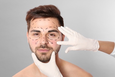 Doctor examining man's face with marker lines for plastic surgery operation on grey background