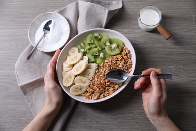 Photo of Woman eating tasty granola with banana and kiwi at grey wooden table, closeup