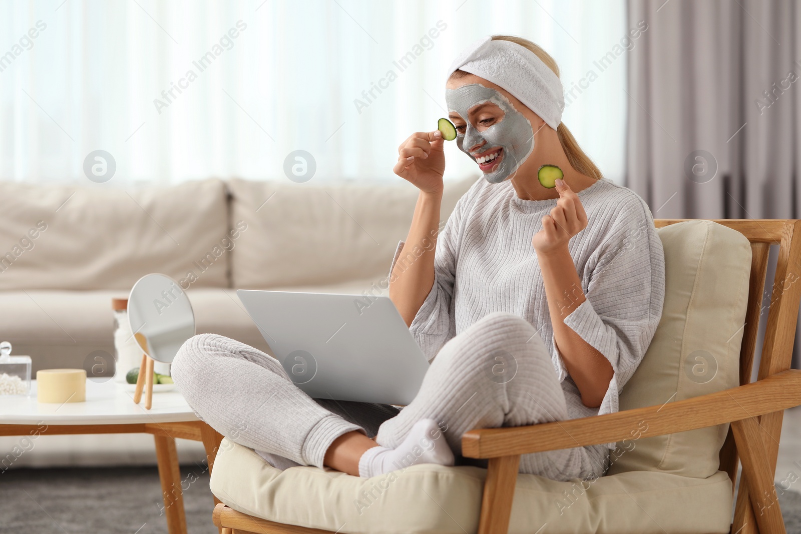 Photo of Young woman with face mask and cucumber slices using laptop at home. Spa treatments