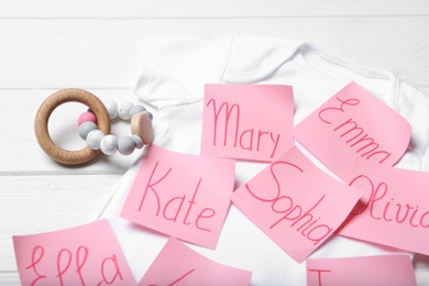 Paper notes with different baby names, child's bodysuit and toy on white wooden table, closeup