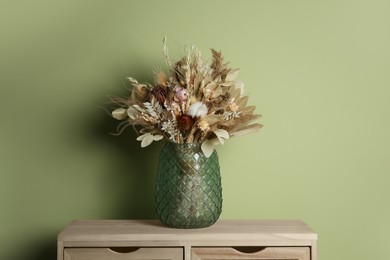 Beautiful dried flower bouquet in glass vase on wooden table near olive wall