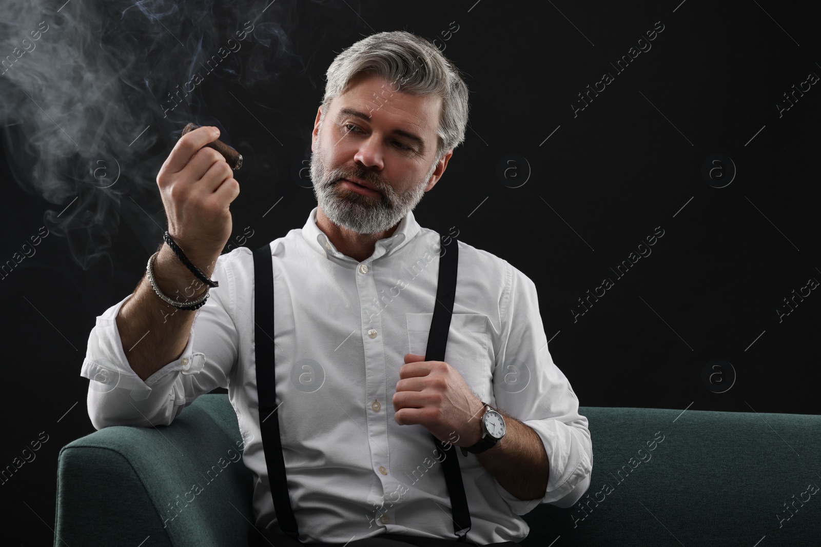 Photo of Bearded man smoking cigar on sofa against black background. Space for text