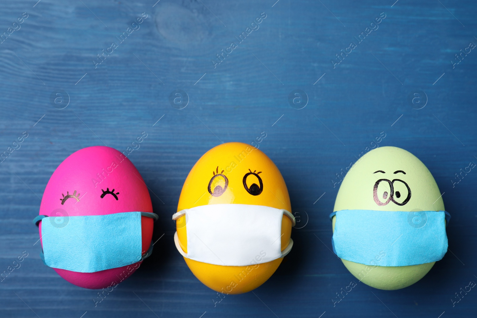 Photo of Bright  eggs in protective masks on blue wooden table, flat lay with space for text. Easter holiday during coronavirus quarantine