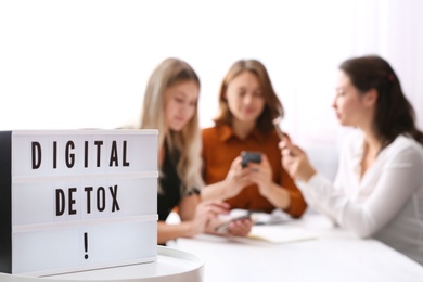 Photo of Women using smartphones at table in office, focus on lightbox with phrase DIGITAL DETOX
