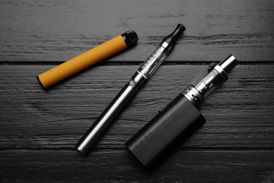 Photo of Different electronic cigarettes on black wooden table, flat lay. Smoking alternative