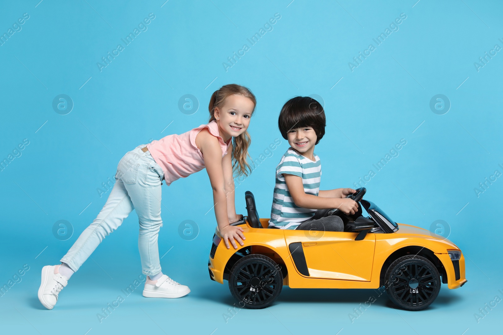 Photo of Cute girl pushing children's electric toy car with little boy on light blue background