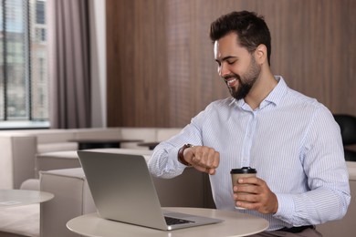 Happy young man checking time while working on laptop at table in office. Space for text