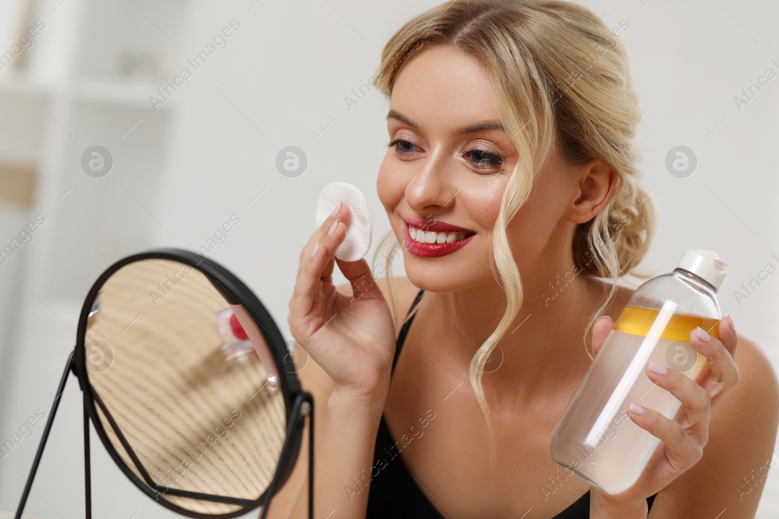 Photo of Smiling woman removing makeup with cotton pad in front of mirror indoors