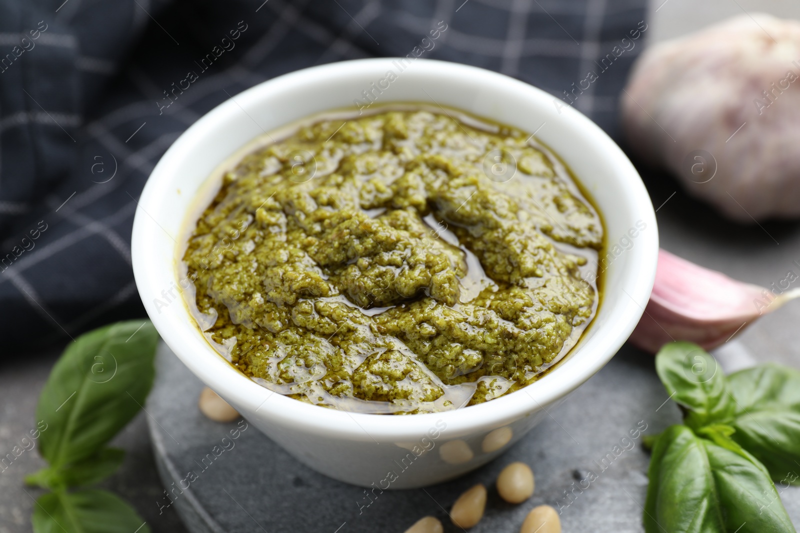 Photo of Tasty pesto sauce in bowl, basil, pine nuts and garlic on table, closeup