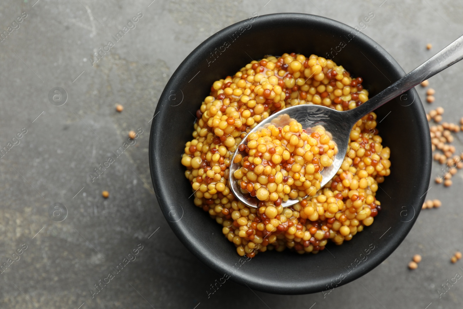 Photo of Whole grain mustard in bowl and spoon on grey table, top view. Space for text